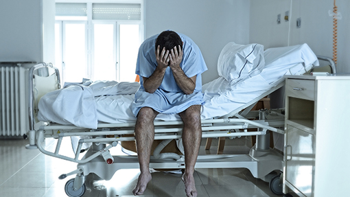 A photograph of a man seated with his legs over the side of a hospital bed in a gown with his head buried in his hands.