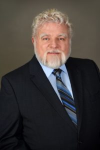A photograph of Dr. John Torwbridge in a dark suit against a brown background smiling.